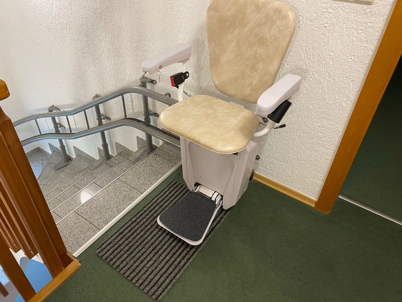 Alpha Treppenlift mit Wandmontage Treppenlift mit beigem Sitz, am Fuß einer Treppe platziert.